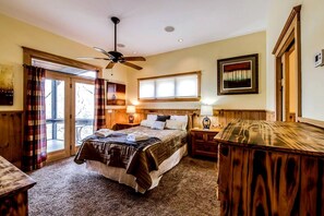 A bedroom with a bed, a wooden desk, a ceiling fan, a window with blinds, a door, and a wall-mounted picture.