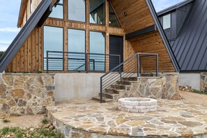 A modern building with a stone foundation, a wooden deck, and a circular stone fire pit. - Family-Friendly A-frame with Outdoor Camp Fire in Texas (New Braunfels)