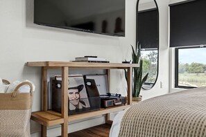 A bedroom with a bed, a TV mounted on the wall, a wooden shelf with books and a picture frame, a plant, and a window with a view.
