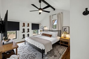 A bedroom with a bed, a desk, a TV, a ceiling fan, and boots on the wall.