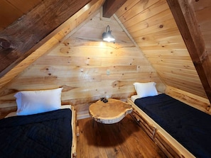 Ein gemütliches Schlafzimmer im Dachgeschoss mit Holzwänden und schräger Decke, ausgestattet mit zwei Betten, einem kleinen runden Tisch und einer einzelnen Lampe.
