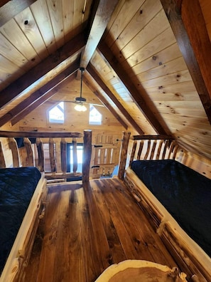 Quarto tipo loft com estrutura de madeira, teto inclinado, duas janelas e uma viga de madeira central.