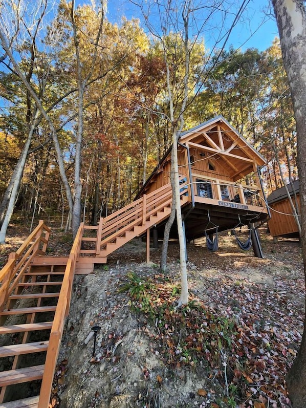 A wooden cabin with a staircase leading up to a deck, surrounded by trees.