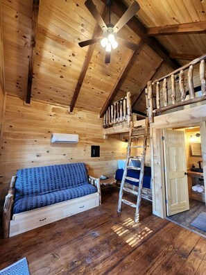 L’intérieur d’une cabane en bois, avec un lit en mezzanine, un canapé, une échelle et un ventilateur de plafond.