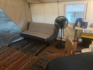 Een tent van binnen, met een bank, een ventilator, een klein tafeltje met spullen en een raam.