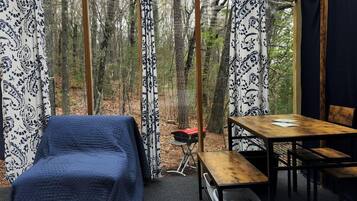 A cozy, enclosed outdoor space with a bed, a table, and a chair, surrounded by patterned curtains and a view of a forest.