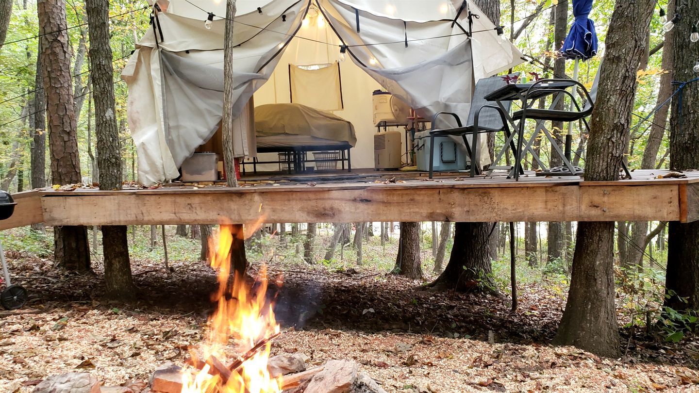 Un campamento con tiendas de campaña, una cama, una mesa y sillas, rodeado de árboles.