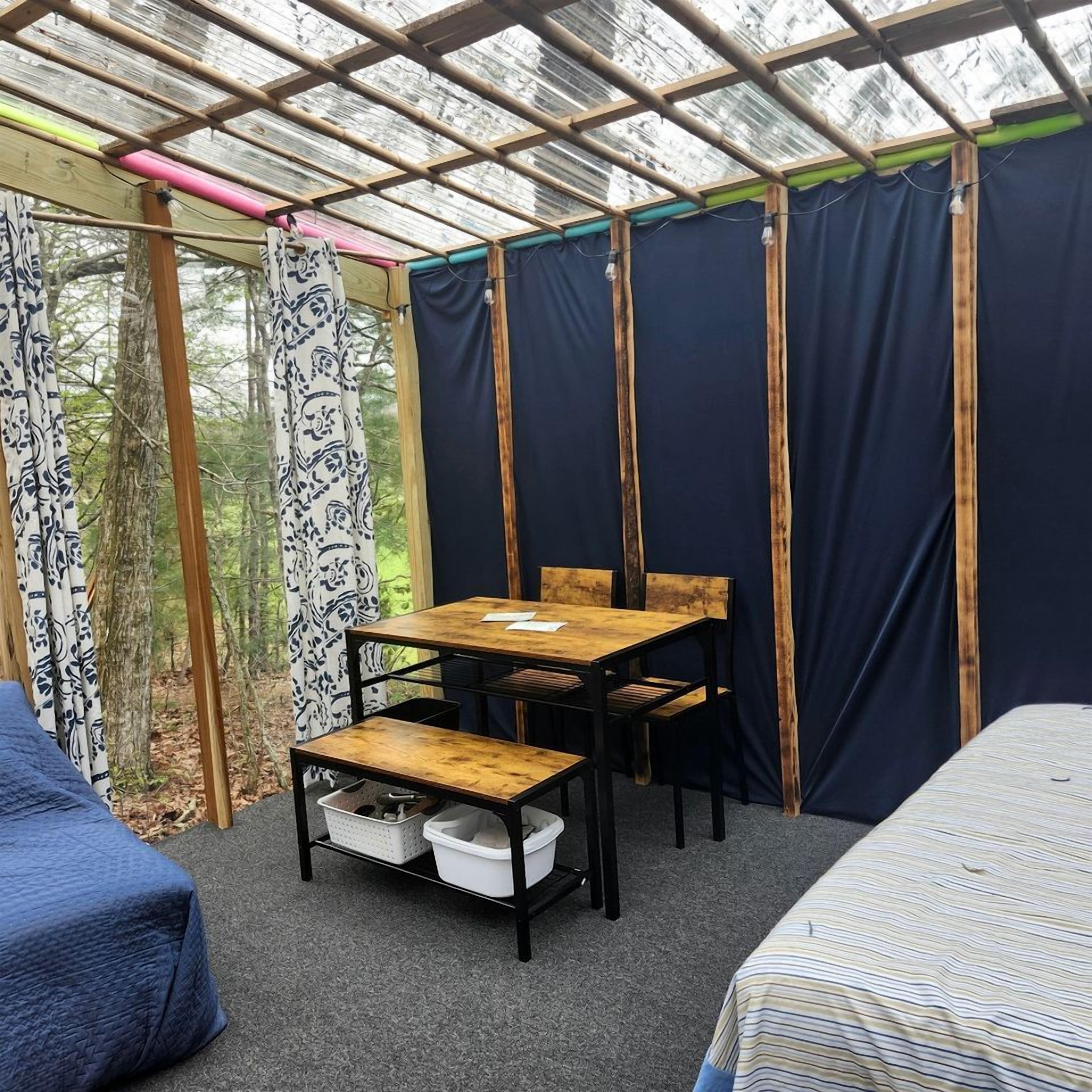 Ein Zimmer mit Holztisch und -stühlen, einem Bett und einem Fenster mit Vorhängen.