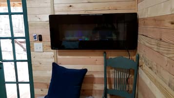 A room with a television mounted on the wall, a wooden chair, a blue cushion, and a window with a view of the outside.