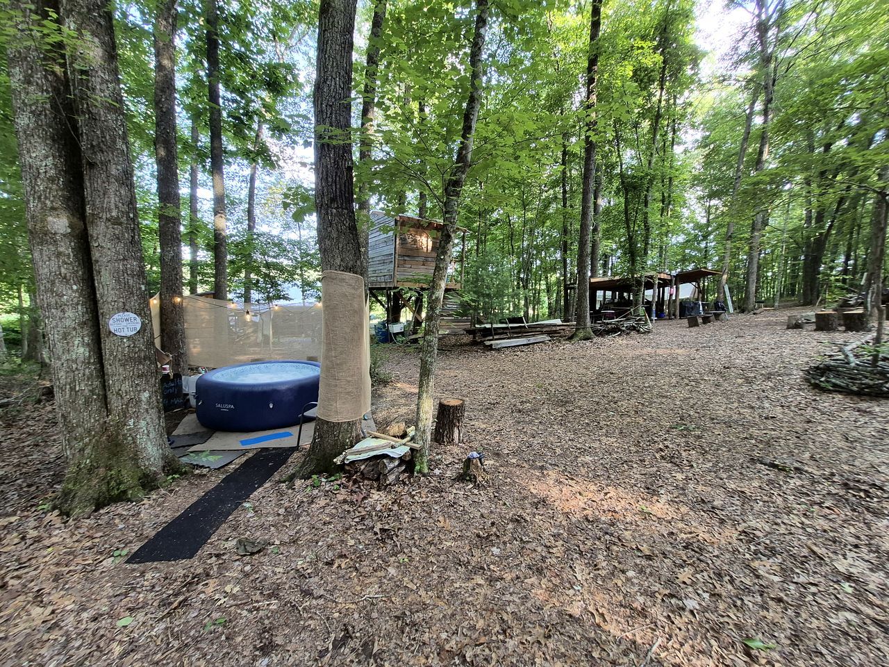 Un terrain de camping boisé, doté d’un abri avec douche, d’un matelas gonflable bleu et de structures en bois.