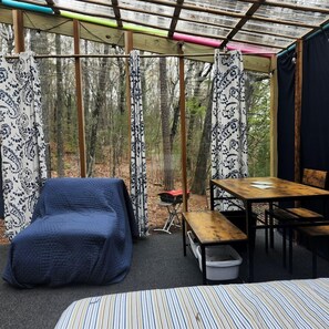 Un espacio exterior íntimo y cerrado, con una cama, una mesa y una silla, rodeado de cortinas con diseños y vistas a un bosque.