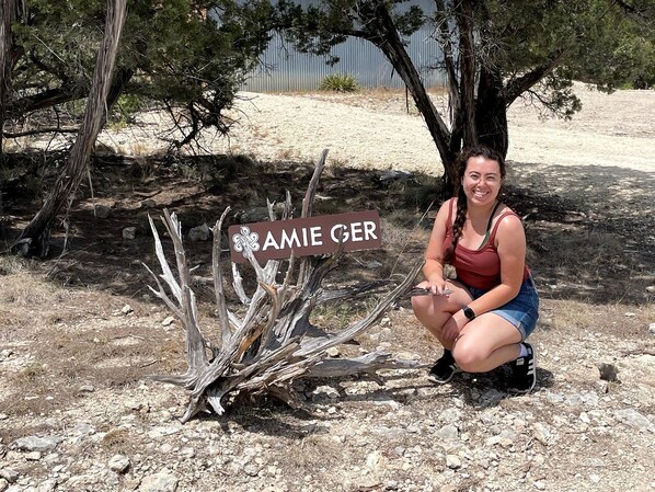 A woman squatting next to a sign with the name 'AMIE GER' in a natural outdoor setting with trees and rocky ground. - Luxurious Romantic Yurt with Hot Tub in Dripping Springs, Texas (Dripping Springs)