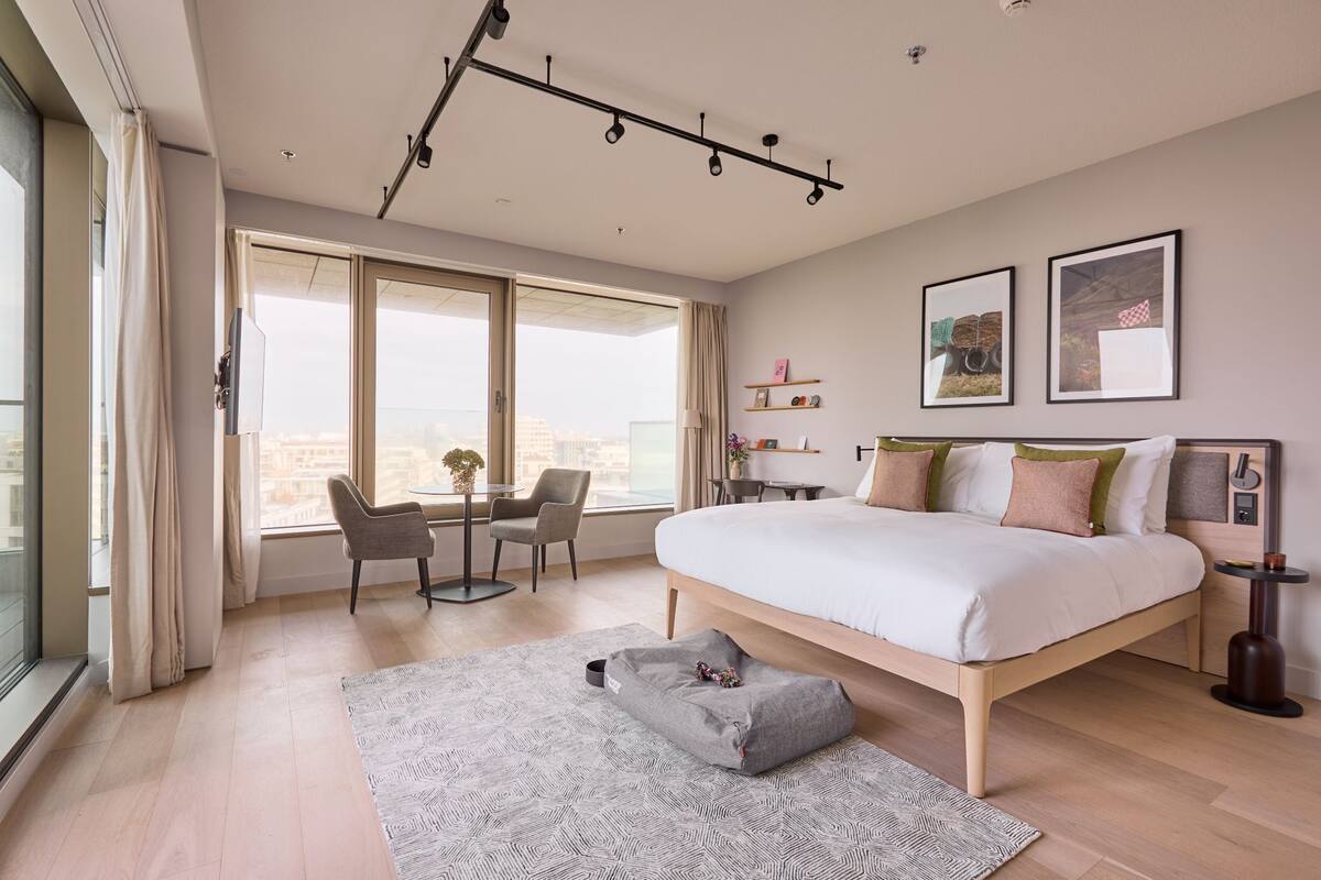 Een moderne slaapkamer met een groot bed, een bureau en een stoel. Er zijn grote ramen en uitzicht op de stad.