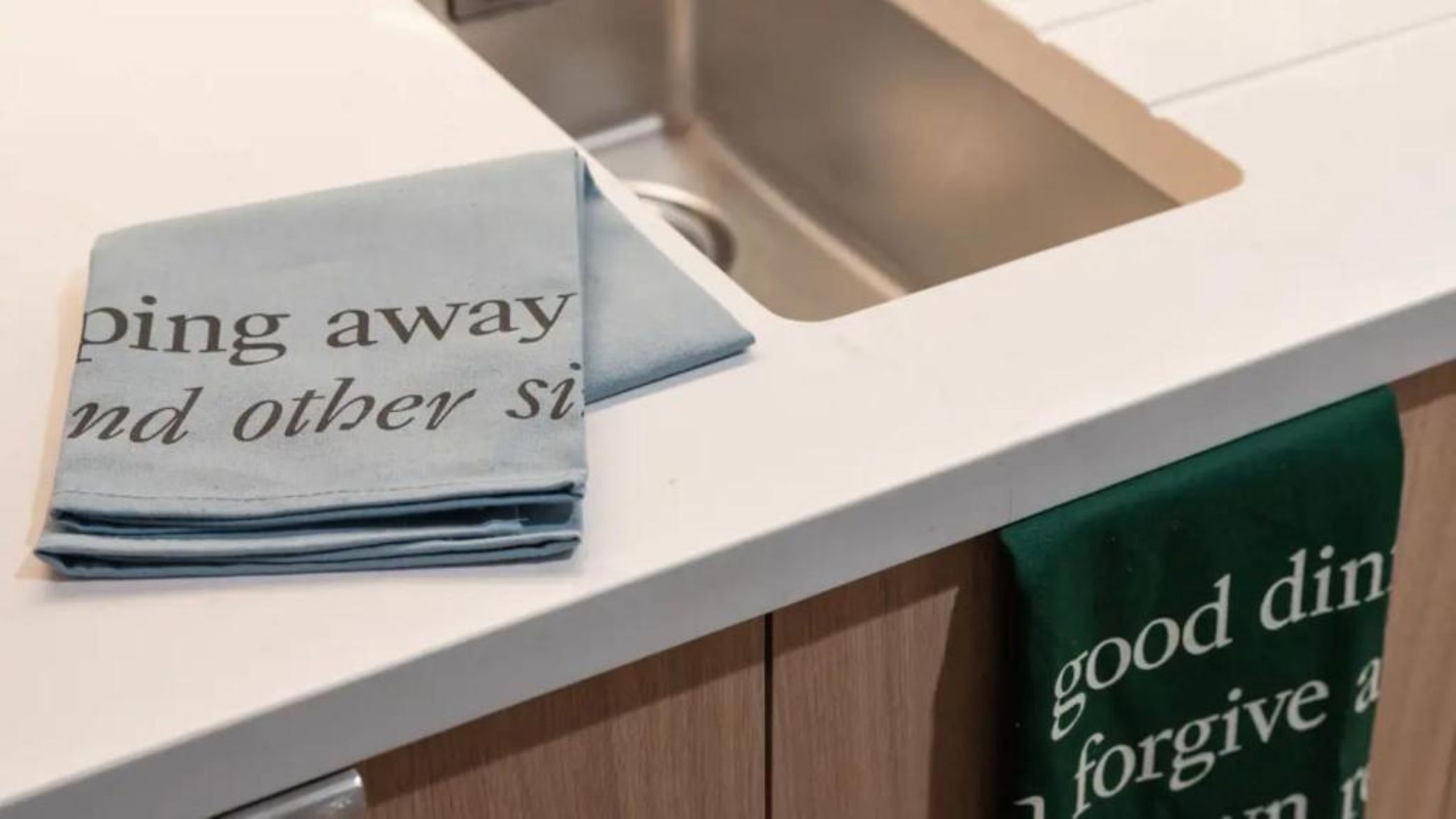 Una encimera de cocina con una toalla que dice “ping away y otras sí” y una toalla verde con el texto “buena comida perdona a”. 