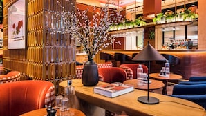 Un intérieur moderne avec un comptoir d’accueil en bois, des sièges rouges et bleus, un vase décoratif orné de branches et un espace bar agrémenté de plantes et d’étagères.