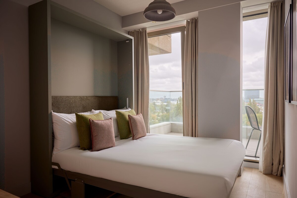 Une chambre d’hôtel moderne dotée d’un grand lit, offrant une vue sur la ville par une fenêtre et comprenant un balcon avec un fauteuil.