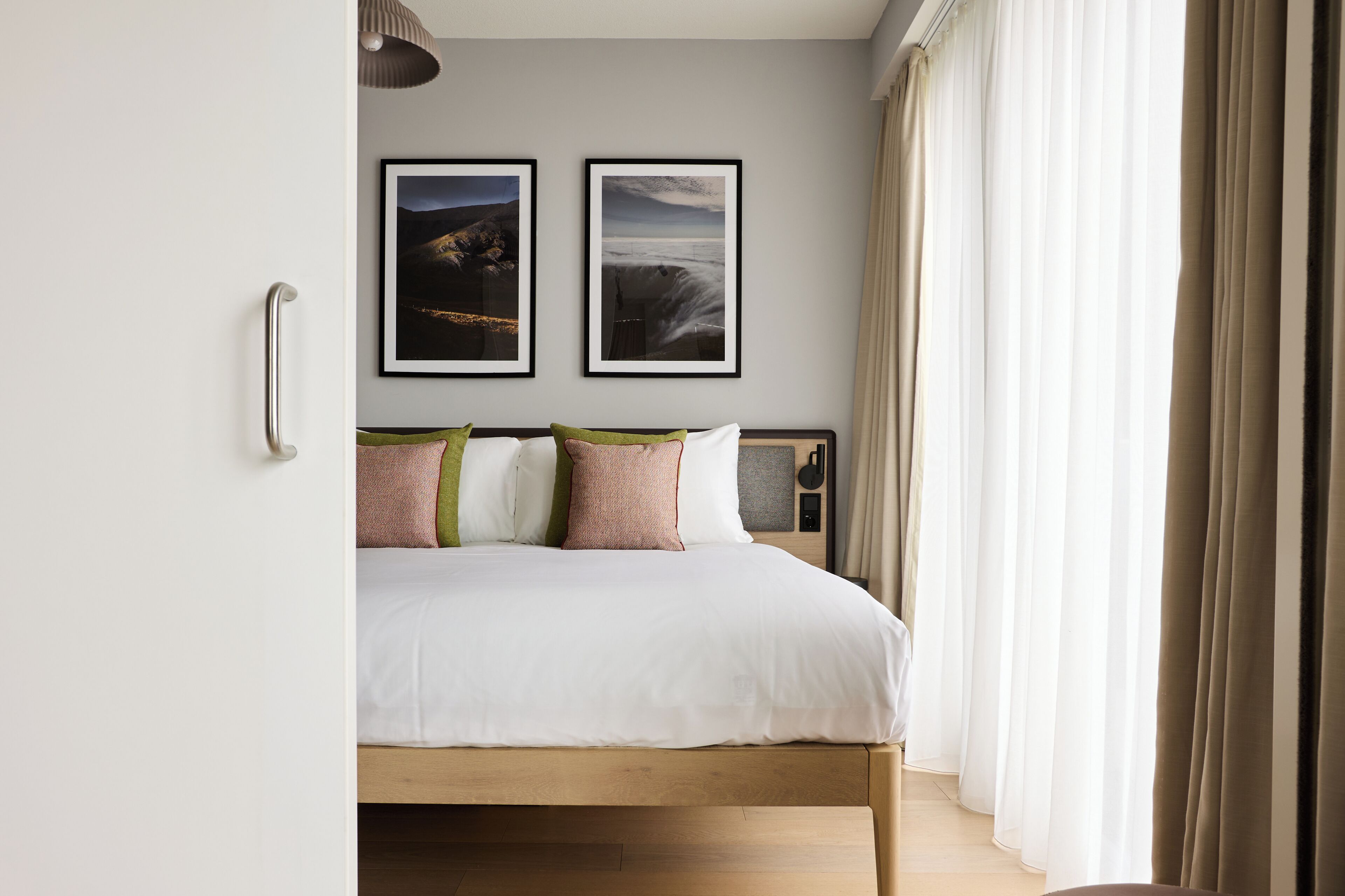 A bedroom with a wooden bed, white bedding, two framed pictures on the wall, a bedside table with a lamp, and sheer curtains.