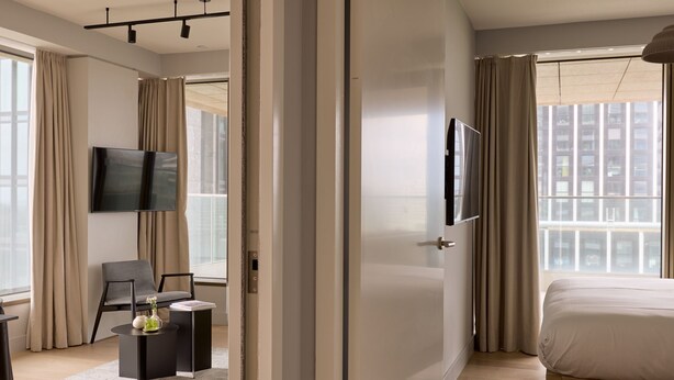 Une chambre d’hôtel moderne dotée d’un grand lit, d’un téléviseur à écran plat fixé au mur, d’un coin salon avec un fauteuil et des tables d’appoint, et de grandes fenêtres avec des rideaux.