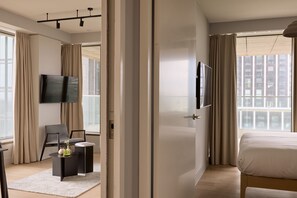 Een moderne hotelkamer met een groot bed, een flatscreen-televisie aan de muur, een zithoek met een fauteuil en bijzettafels, en grote ramen voorzien van gordijnen.