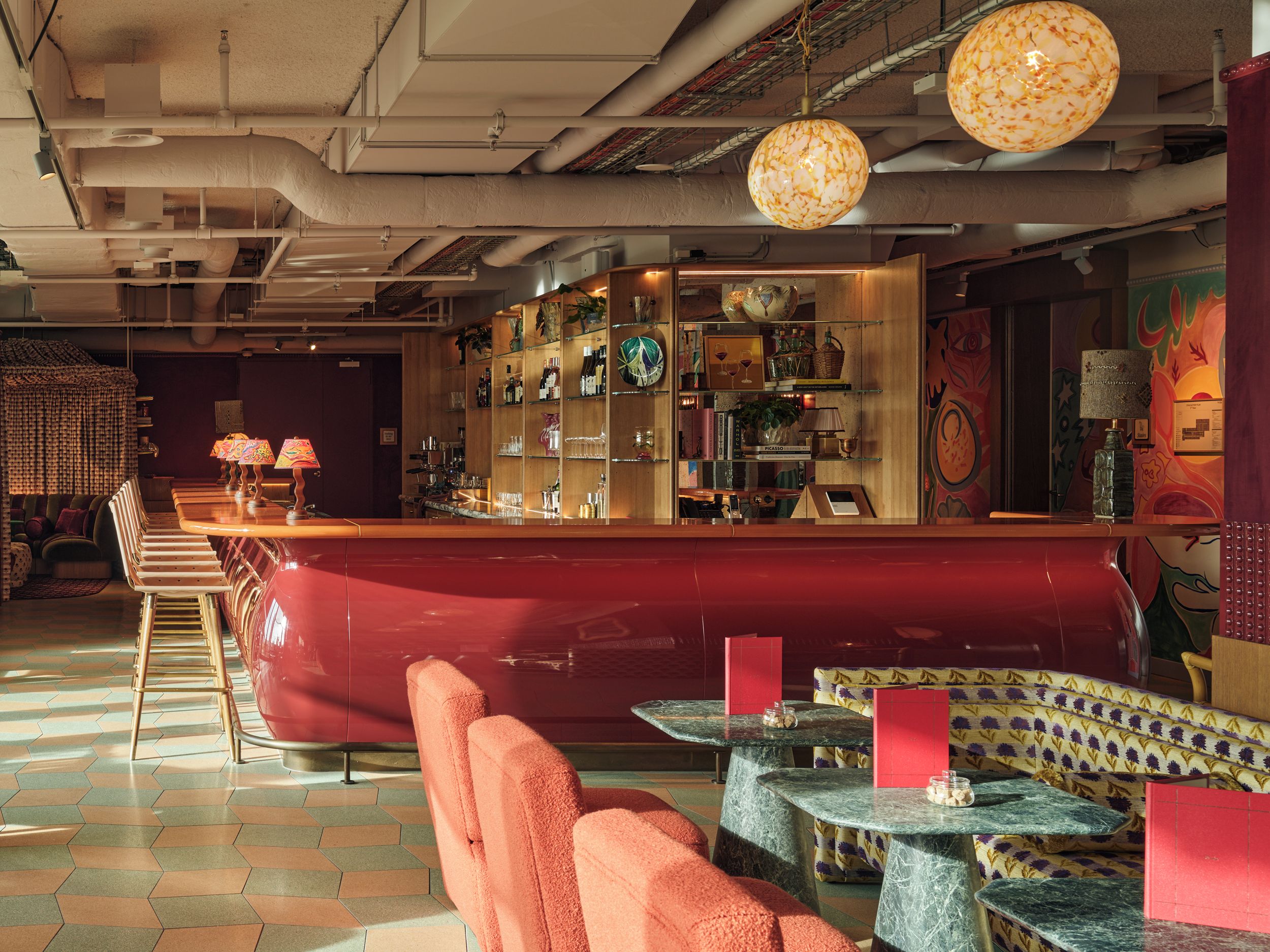 Een bar met een rode toonbank, zitplaatsen met een patroon bekleding, en diverse flessen en glazen op planken.