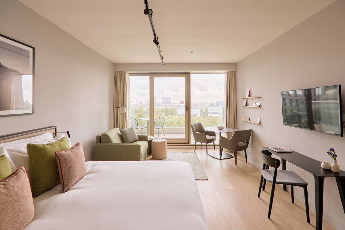 Een hotelkamer met een groot bed, een bureau, een stoel, een bank en uitzicht op de stad door grote ramen.