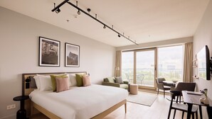 Une chambre d’hôtel moderne dotée d’un grand lit, d’un coin salon avec une table et des chaises, et d’un balcon offrant une vue sur la ville.