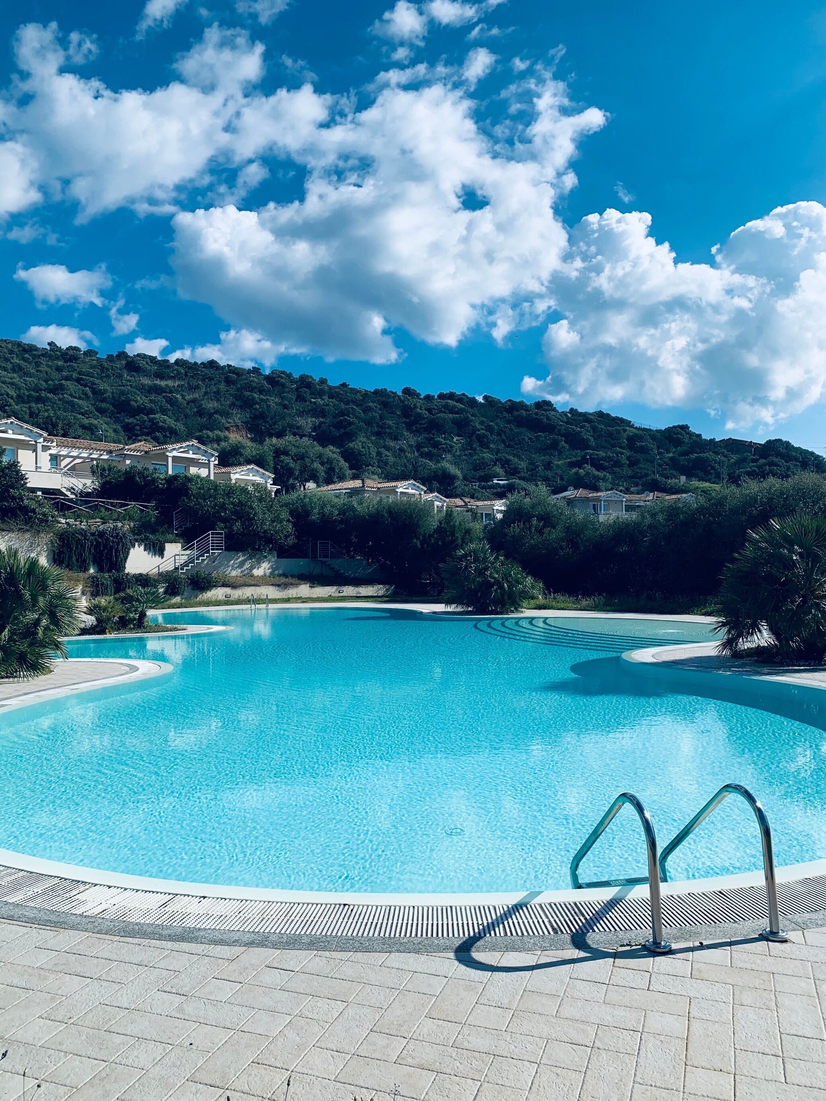 A clear blue swimming pool with a tiled deck, surrounded by greenery and a hilly landscape.
