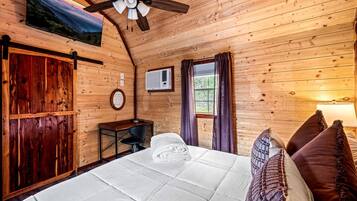 A wooden cabin room with a bed, a desk, a chair, a ceiling fan, and a window with curtains.