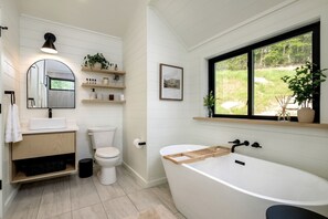 A modern bathroom with a freestanding bathtub, a toilet, a sink, and a window with a view of greenery.