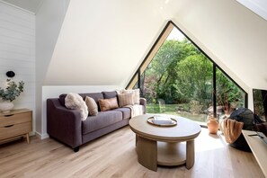 A modern loft bedroom with a sloped ceiling, a dark-colored sofa, a round wooden table, and a large window offering a view of a garden.
