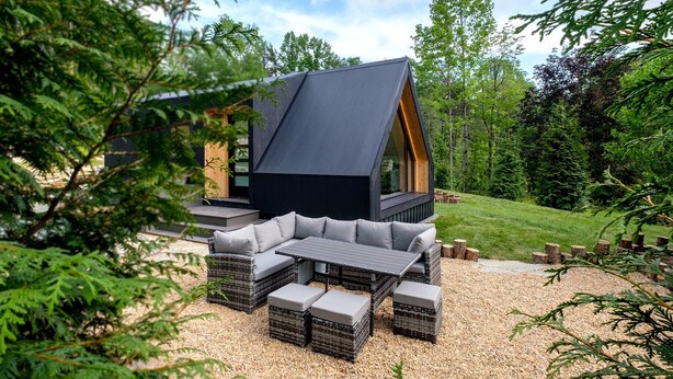 Uma cabana moderna com telhado inclinado, área de estar externa e vista para uma paisagem arborizada.