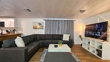 Un salon moderne avec un canapé d’angle, une table basse, un téléviseur et, au fond, un coin cuisine.