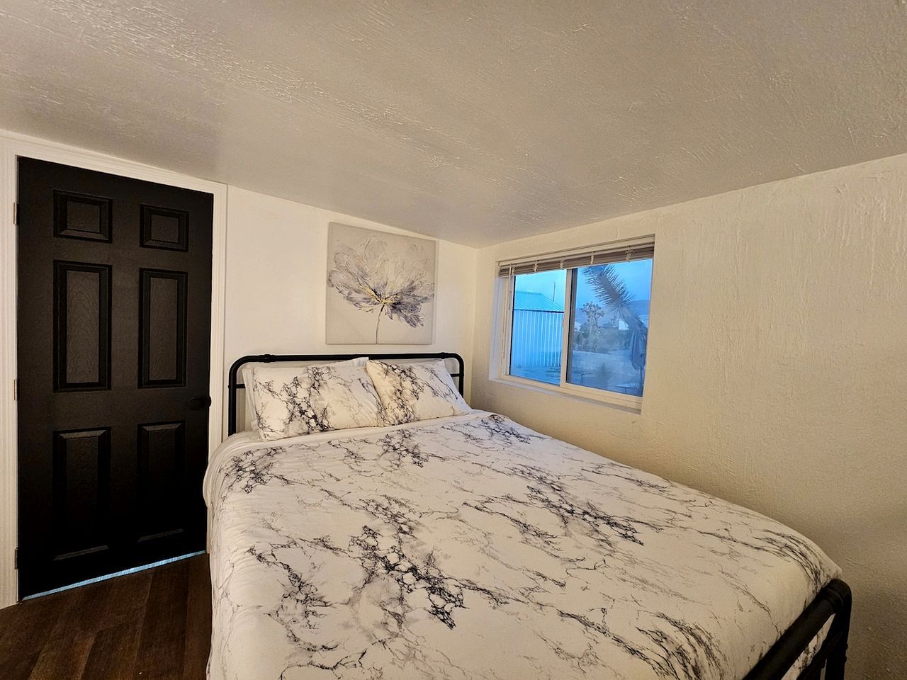 A bedroom with a bed, a window, a door, and a painting on the wall.