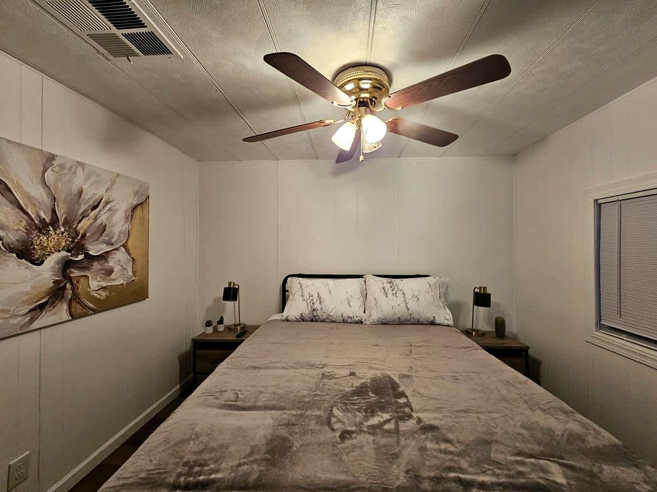 Ein Schlafzimmer mit einem Bett, einem Deckenventilator, einem Nachttisch, einer Lampe und einem Wandbild.