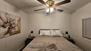 Ein Schlafzimmer mit einem Bett, einem Deckenventilator, einem Nachttisch, einer Lampe und einem Wandbild.