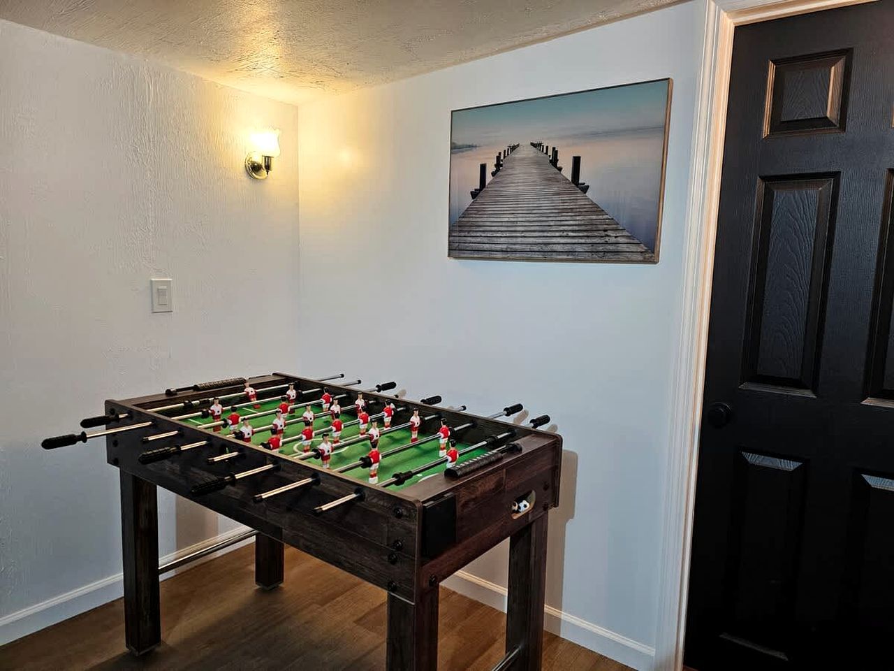Un tavolo da calcio in una stanza con una porta, una luce a parete e un quadro raffigurante un molo.