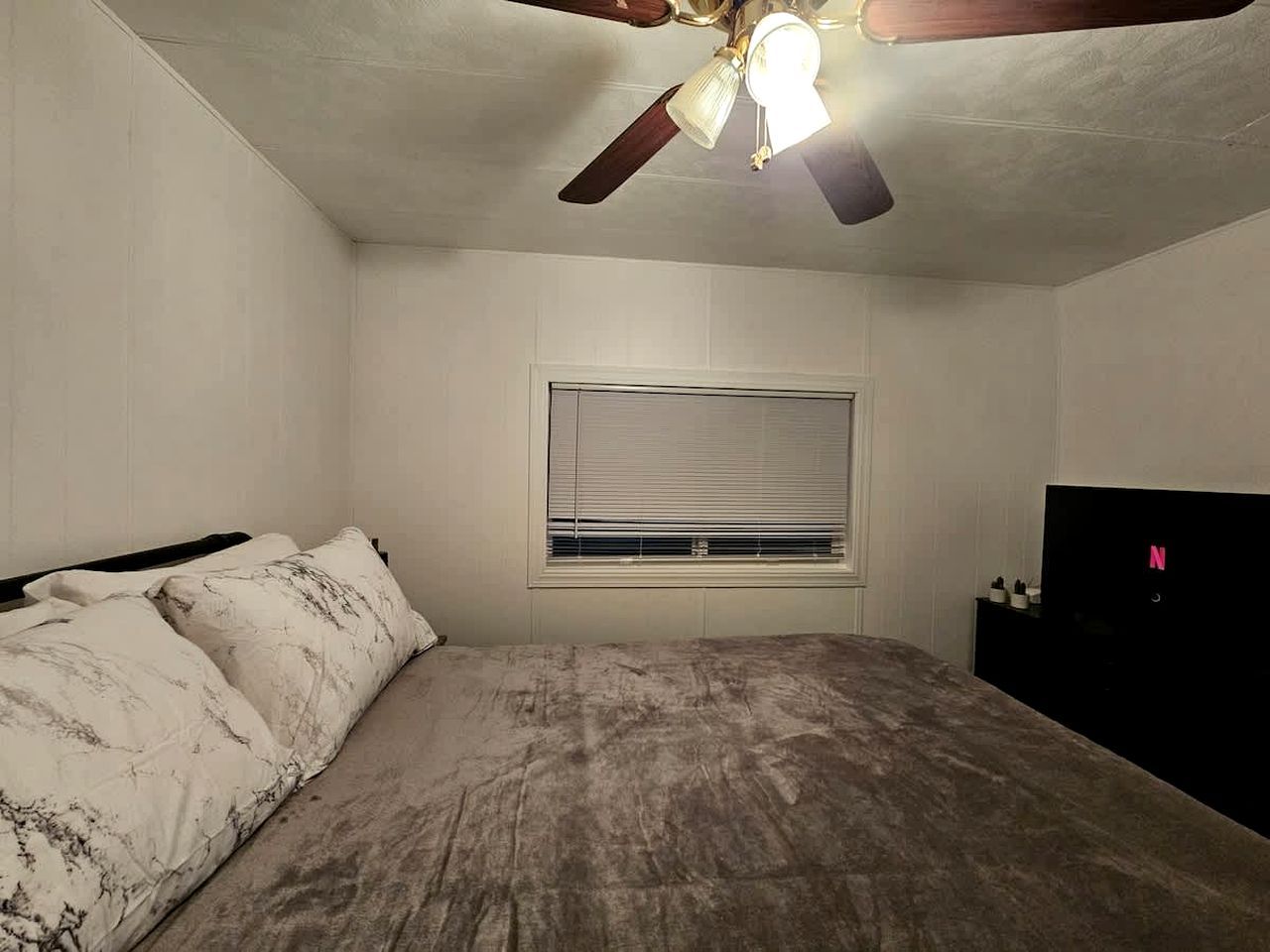 Une chambre à coucher avec un lit, un ventilateur de plafond, une fenêtre avec des stores et un téléviseur posé sur un meuble.