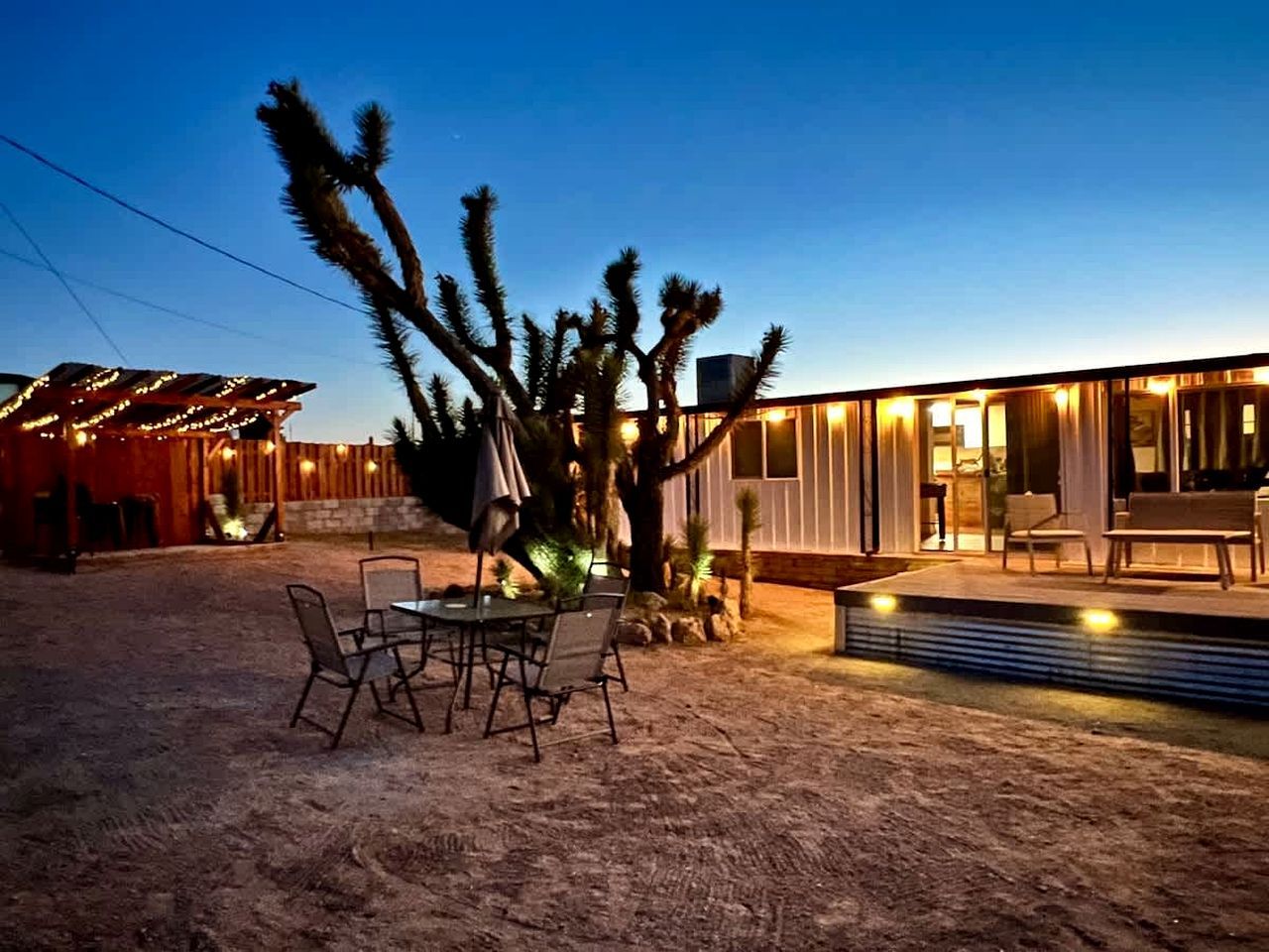 Un espace extérieur aménagé dans un style désertique, avec une table et des chaises, un cactus et un bâtiment moderne doté de grandes fenêtres.