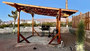 A wooden pergola with string lights, a barbecue grill, and a small plant in a backyard with a dirt floor and a wooden fence.