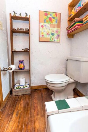 A bathroom with a toilet, wooden shelves with decorative items, a framed floral artwork, and a wooden floor. - Rustic Cabin Rental Perfect for Families in Texas Hill County (Marble Falls)