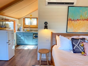 A compact room with a bed, a stool, a small table, a kitchenette, and a window.