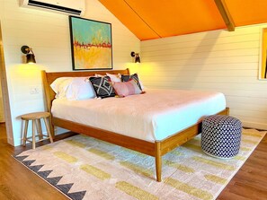 Un dormitorio con una cama de madera, un taburete, una alfombra estampada y una lámpara fijada a la pared.