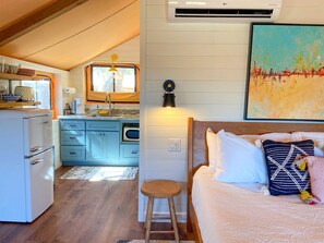 A compact room with a bed, a stool, a small table, a kitchenette, and a window.