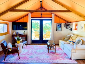 A cozy living room with a TV, comfortable seating, a fan, and a view through a large door.