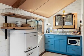 Une cuisine compacte avec un réfrigérateur Galanz blanc, des placards bleus, une étagère en bois où sont posées des tasses et une fenêtre donnant sur un paysage agréable.