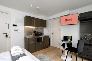 Ein modernes Hotelzimmer mit Küchenzeile, einem Bett mit ordentlich gefalteten Handtüchern, einem kleinen Tisch mit Pflanze und einem Schild mit der Aufschrift 'GUEST READY'.