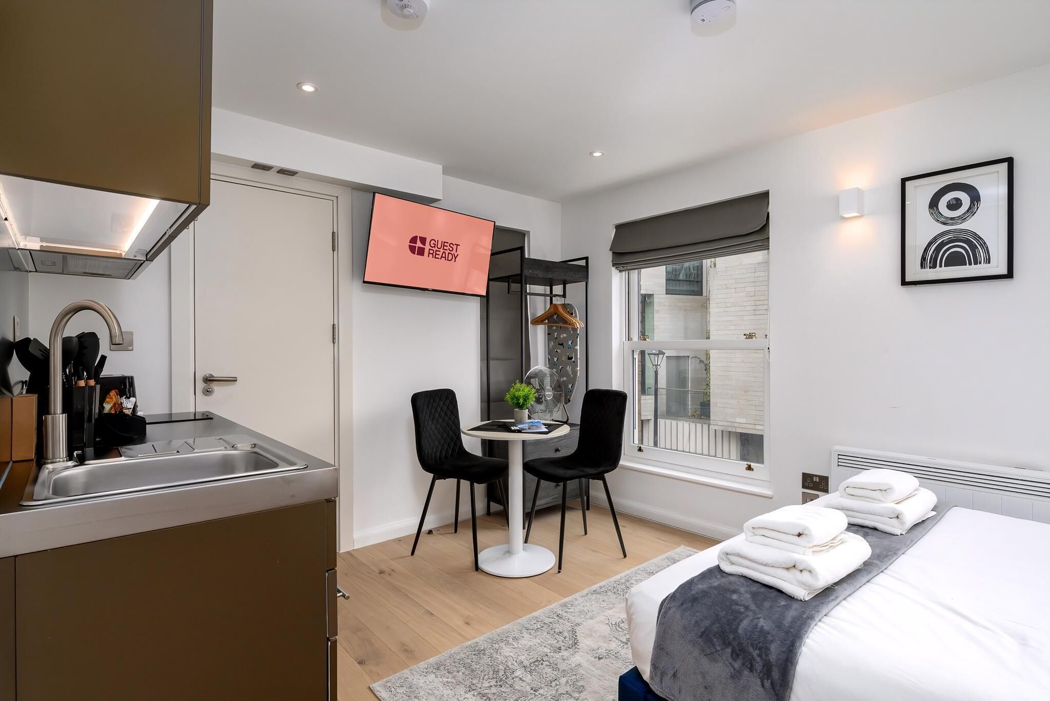 A modern hotel room with a bed, a small dining table with chairs, a kitchen area, and a TV displaying "GUEST READY".