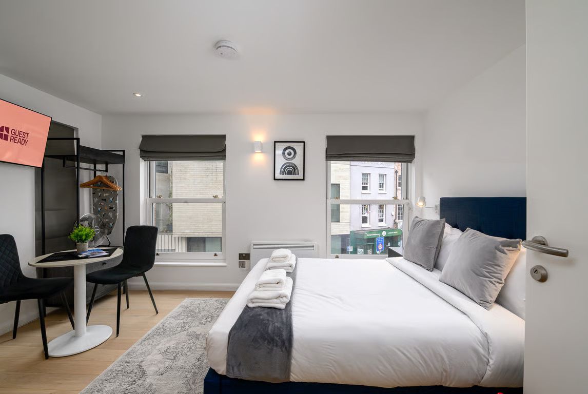 A modern hotel room with a bed, a desk, and a chair. There is a window with a view of buildings outside.