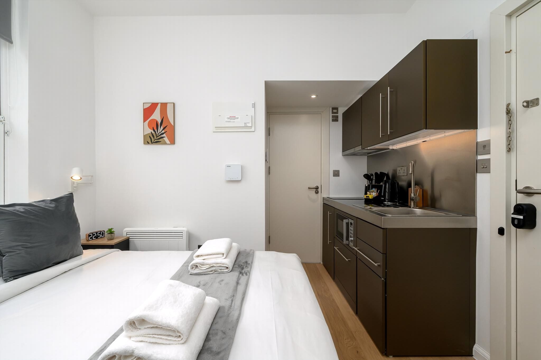 Een compacte hotelkamer met een bed, een klein nachtkastje met een lamp, een wandairconditioner, een kitchenette met kastjes en een kookplaat, en een deur die naar een andere kamer leidt.