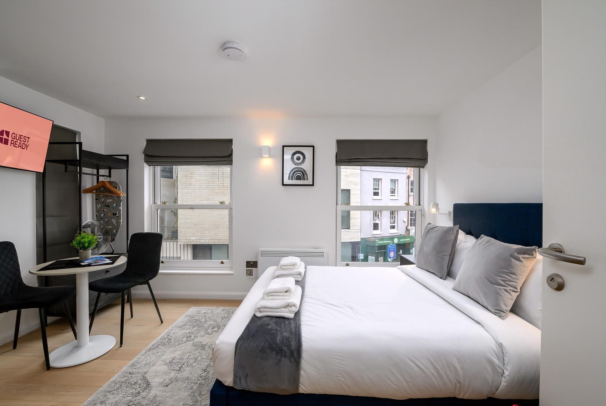 A modern hotel room with a bed, a desk, and a chair. There is a window with a view of buildings outside.